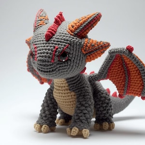 以下が含まれることがあります： グレー、オレンジ、赤の色合いの、かぎ針編みのドラゴンフィギュア。手作りの生き物は大きな目、翼、角を持っています。ドラゴンは四つん這いになっており、その詳細な職人技を披露しています。