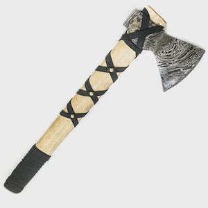 May include: A handcrafted axe with a patterned steel blade and a light-colored wooden handle. The handle is wrapped with black leather straps and accented with gold-colored studs. The bottom of the handle is wrapped in black rubber.