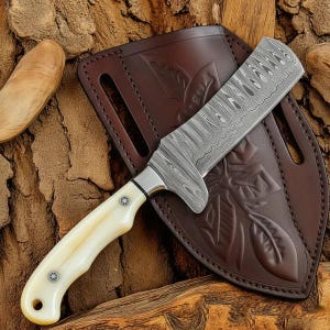 May include: A fixed-blade knife with a patterned Damascus steel blade and a smooth, off-white handle secured by two rivets. The knife is positioned over a dark brown leather sheath featuring an embossed leaf design and two belt slots.