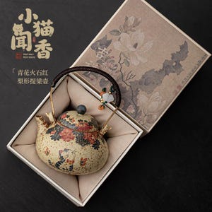 Puede incluir: Una tetera de cerámica con un diseño floral y de gato, presentada en una caja decorativa. La tetera tiene un asa marrón y una tapa con un pomo azul oscuro. La caja presenta un patrón floral y texto chino.