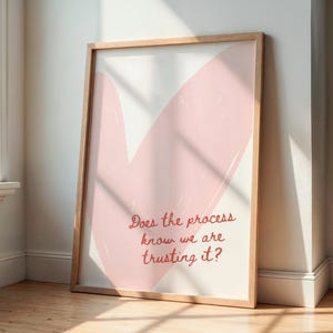 Op de afbeelding: Een ingelijste kunstdruk met een licht houten frame. Het kunstwerk toont een grote, abstracte roze penseelstreek op een witte achtergrond. De tekst "Does the process know we are trusting it?" is in cursief geschreven.