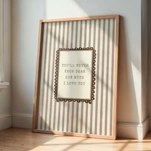 Puede incluir: Una impresión enmarcada con un fondo a rayas y un diseño rectangular central. El texto "YOU'LL NEVER KNOW DEAR HOW MUCH I LOVE YOU" se muestra en una fuente decorativa. El marco es de madera.