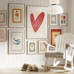 May include: A gallery wall featuring framed art prints with various designs, including a red heart, whales, and a giraffe. A white rocking chair and an open book are in the foreground. The wall is painted white.