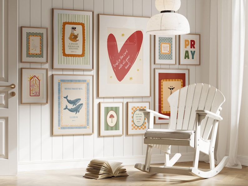 May include: A gallery wall featuring framed art prints with various designs, including a red heart, whales, and a giraffe. A white rocking chair and an open book are in the foreground. The wall is painted white.
