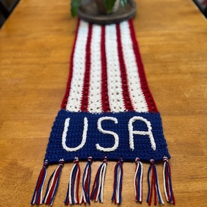 May include: A red, white, and blue crocheted table runner. The runner has red and white stripes, with a blue section at the end spelling "USA" in white. The bottom edge is finished with red, white, and blue tassels.