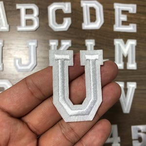 Puede incluir: Un parche bordado blanco y gris con la letra "U" se sostiene en una mano, con otros parches de letras en el fondo. Los parches tienen un estilo universitario con un borde blanco y relleno gris.