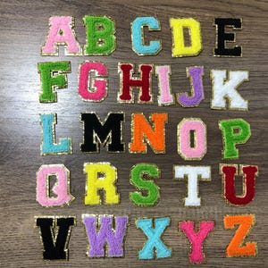 Könnte beinhalten: Eine Sammlung farbenfroher, gestickter Stoffbuchstaben, die jeweils goldfarben umrandet sind. Die Buchstaben sind in verschiedenen Farben gehalten, darunter Rosa, Grün, Blau, Gelb, Schwarz, Rot, Lila und Weiß. Die Buchstaben sind auf einer Holzoberfläche angeordnet.