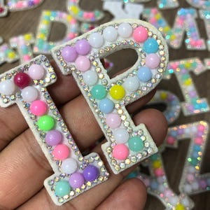 May include: Close-up of the letter "I" and "P" patches. The white letters are decorated with colorful beads and surrounded by rhinestones. Other letters are visible in the background.