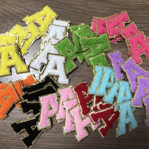 May include: A collection of colorful, embroidered letter "A" patches. The patches are in various colors, including yellow, white, green, pink, purple, orange, black, light blue, and red. Each letter is outlined with gold trim.