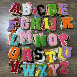 May include: A collection of colorful, embroidered fabric letter patches. Each letter is a different color, including red, orange, green, and blue, with gold trim. The letters are in various sizes and fonts.