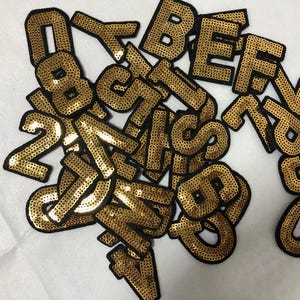 Könnte beinhalten: Eine Ansammlung goldener Buchstaben und Zahlen mit schwarzen Rändern. Die Buchstaben und Zahlen sind auf einer weißen Oberfläche verstreut. Die Pailletten verleihen den Buchstaben ein glänzendes, strukturiertes Aussehen. Ideal zum Basteln.