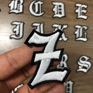 May include: White embroidered number 1 patch with a black border, held in hand. The patch has a gothic font style. Other embroidered letters are visible in the background on a wooden surface.
