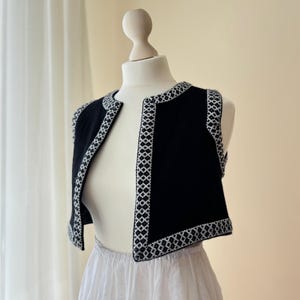 Può includere: Un gilet nero con finiture geometriche bianche è esposto su un manichino. Il gilet ha un design semplice, aperto sul davanti e una lunghezza corta. Il bordo bianco crea un contrasto sorprendente con il tessuto nero.