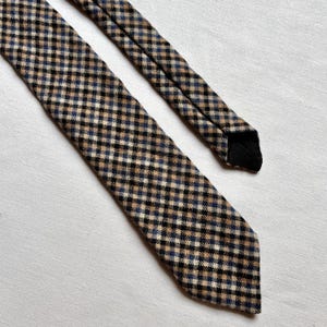 May include: A checkered tie in a classic pattern. The tie features a repeating pattern of black, tan, and blue squares. The tie is made of a textured fabric and has a black lining on the back.