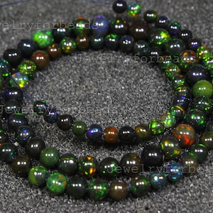 May include: A strand of black opal beads, displaying iridescent colours of green, blue, and orange. The round beads are strung together, creating a shimmering effect. The beads are on a dark grey surface.