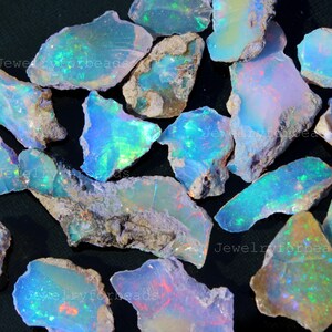 May include: A collection of raw opal stones with vibrant blue, green, and iridescent flashes. The rough-cut gemstones have a natural, earthy appearance, with a mix of light and dark areas. The image includes the text "Jewelryforbeads".