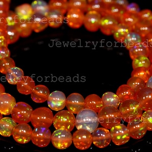 May include: Strands of orange opal beads with iridescent flashes of color. The beads are round and vary in size, strung together to create a necklace or bracelet. The beads have a glossy finish and are set against a black background.