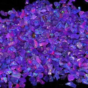 Könnte beinhalten: Ein Haufen schillernder Opal-Edelsteine in Lila-, Blau- und Rosatönen. Die grob geschnittenen Steine sind auf einer dunklen Oberfläche verstreut und erzeugen eine funkelnde, farbenfrohe Darstellung. Die Opale variieren in Größe und Form.