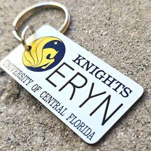 Peut inclure: Un porte-clés rectangulaire avec un anneau argenté. Le fond blanc porte les mots "KNIGHTS" et "ERYN" en noir, avec le logo de l'Université de Central Florida en jaune et bleu.