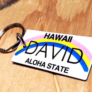 Op de afbeelding: Een rechthoekige sleutelhanger met een zilveren ring. De sleutelhanger heeft de naam "DAVID" in grote zwarte letters, met "HAWAII" erboven en "ALOHA STATE" eronder. Een regenboogontwerp loopt over de bovenkant.