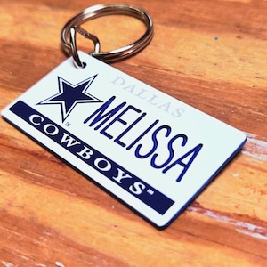 May include: A rectangular keychain with a silver ring. The keychain has a white background with the name "MELISSA" in blue, the word "DALLAS" above, and "COWBOYS" below. A blue star logo is on the top left.