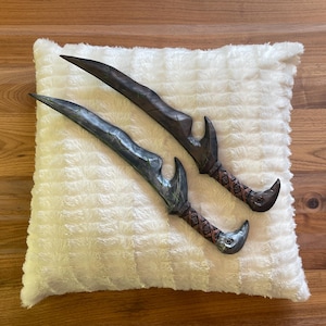 May include: Two decorative daggers with dark gray blades and brown handles rest on a fluffy white pillow. The handles have a woven pattern and a curved, bird-like design at the end. The daggers are displayed on a wooden surface.