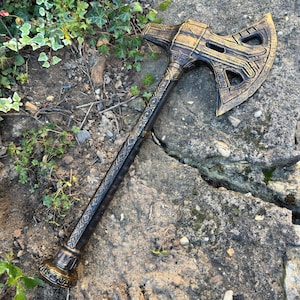 May include: A bronze-colored axe with intricate detailing on the head and handle. The axe head features cut-out designs and a textured surface. The handle has a patterned design. The axe is lying on a stone surface with greenery.