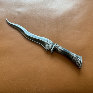 May include: A silver-colored dagger with a curved blade and ornate handle. The handle features detailed scrollwork and a dark finish. The dagger is displayed on a brown leather surface.
