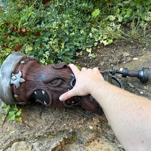 May include: A detailed, brown, cylindrical object with a carved face design and metal accents. The object has a handle and is lying on a stone surface. The object appears to be a decorative piece or prop.