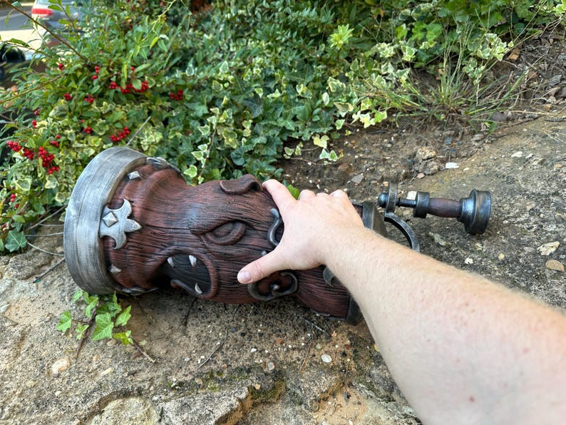 May include: A detailed, brown, cylindrical object with a carved face design and metal accents. The object has a handle and is lying on a stone surface. The object appears to be a decorative piece or prop.