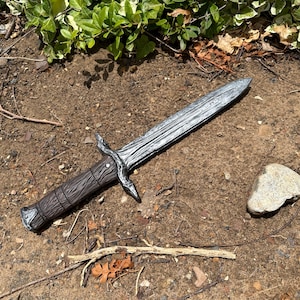 May include: A detailed, silver-colored dagger with a textured blade and a brown handle. The dagger has a decorative crossguard and is lying on the ground. The overall design suggests a fantasy or historical theme.