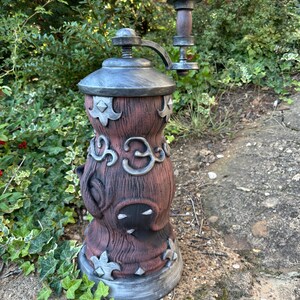 May include: A large, decorative outdoor pepper grinder with a rustic, fantasy design. The grinder is primarily brown with silver accents, featuring ornate details and a crank handle. It stands on a stone surface, surrounded by greenery.