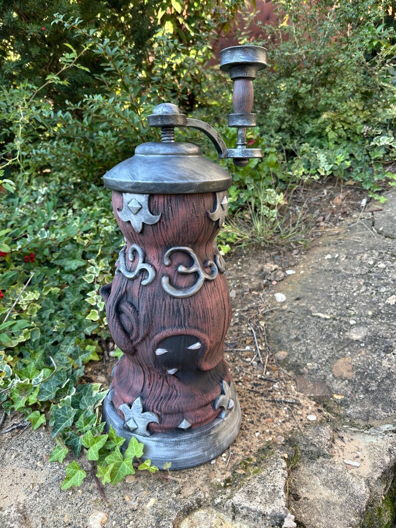 May include: A large, decorative outdoor pepper grinder with a rustic, fantasy design. The grinder is primarily brown with silver accents, featuring ornate details and a crank handle. It stands on a stone surface, surrounded by greenery.