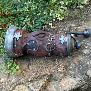 May include: A decorative, fantasy-themed bottle stopper. The stopper is primarily brown with silver accents, featuring a carved design of a creature's face and ornate metalwork. The stopper has a handle and is lying on a stone surface.