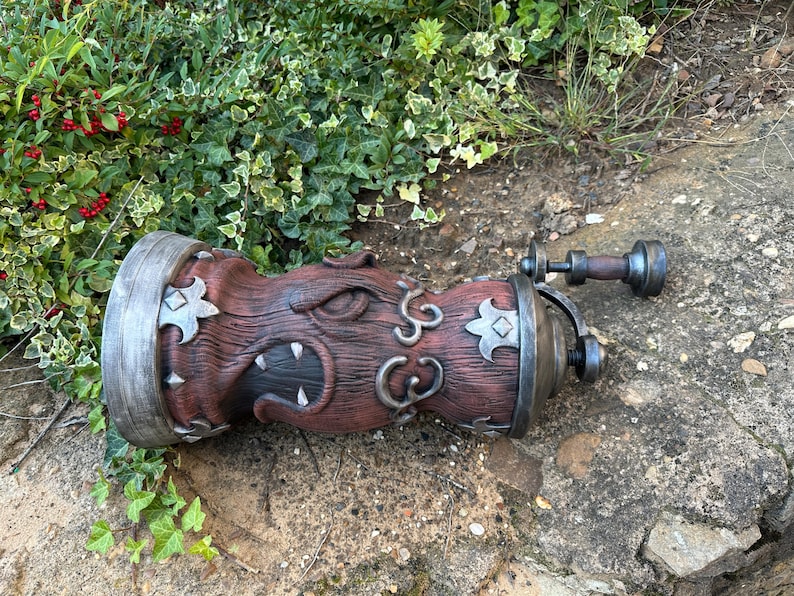 May include: A detailed, brown and gray decorative object with a unique design. The object has a cylindrical body with ornate carvings, metal accents, and a handle. The design includes stylized floral shapes and a textured surface.
