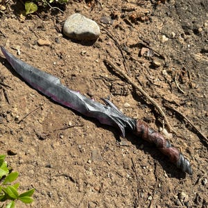 Könnte beinhalten: Ein silberfarbener Fantasy-Dolch mit einem dekorativen Griff, der mit braunem Leder umwickelt ist. Die Klinge hat einen leichten rosa Farbton am Rand. Der Dolch liegt auf einem Bett aus Erde und kleinen Steinen.