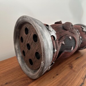 May include: A decorative, cylindrical object with a wooden, perforated end and a textured, reddish-brown body. The object has silver-colored accents and is resting on a wooden surface. The design has a fantasy theme.