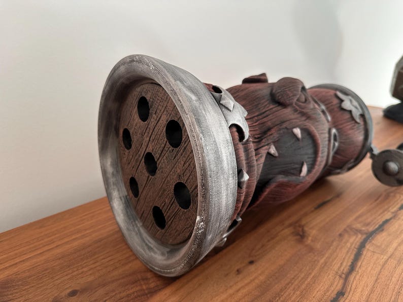 May include: A decorative, cylindrical object with a wooden, perforated end and a textured, reddish-brown body. The object has silver-colored accents and is resting on a wooden surface. The design has a fantasy theme.