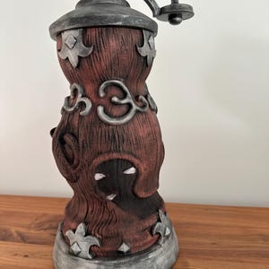 May include: A decorative, antique-style coffee grinder. The grinder features a dark red, textured body with silver-colored accents and a wooden handle. The design includes ornate, stylized floral and geometric shapes.