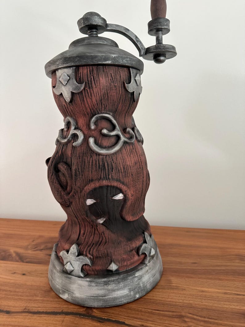 May include: A decorative, antique-style coffee grinder. The grinder features a dark red, textured body with silver-colored accents and a wooden handle. The design includes ornate, stylized floral and geometric shapes.