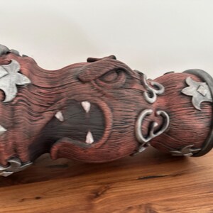 May include: A decorative bottle stopper with a carved, menacing face design. The stopper features a red-brown wooden texture with silver accents and a metal ring. The design includes sharp teeth and stylized metal embellishments.