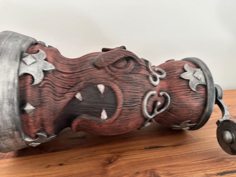 May include: A decorative bottle stopper with a carved, menacing face design. The stopper features a red-brown wooden texture with silver accents and a metal ring. The design includes sharp teeth and stylized metal embellishments.
