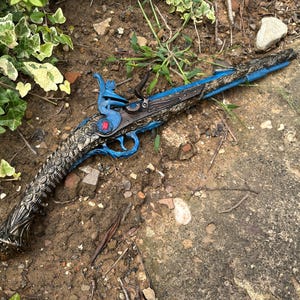 以下が含まれることがあります： 精巧な装飾が施されたアンティーク風のピストル。銃身とグリップは濃い色で、トリガー、グリップ、装飾部分に青色のアクセントがあります。小さな赤いエンブレムが見えます。