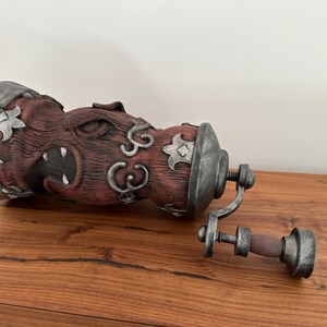May include: A detailed, reddish-brown bottle stopper with a carved monster face design and silver-colored accents. The stopper is separated from its screw mechanism, which is also silver and brown. The item is on a wooden surface.