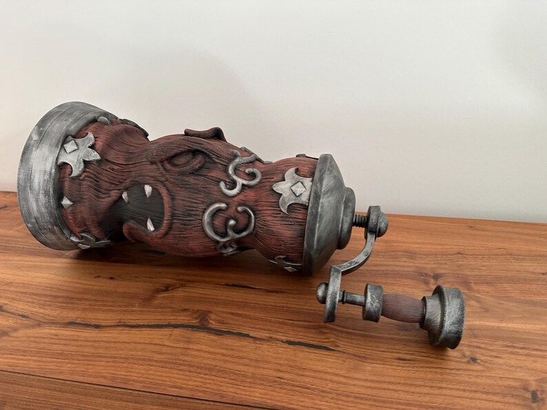 May include: A detailed, reddish-brown bottle stopper with a carved monster face design and silver-colored accents. The stopper is separated from its screw mechanism, which is also silver and brown. The item is on a wooden surface.