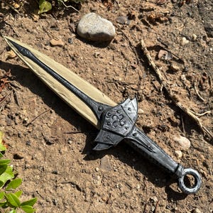 May include: A decorative dagger with a black handle and blade, featuring a silver-colored guard with an ornate design. The blade has a light tan color. The dagger is lying on a bed of dirt and small rocks.