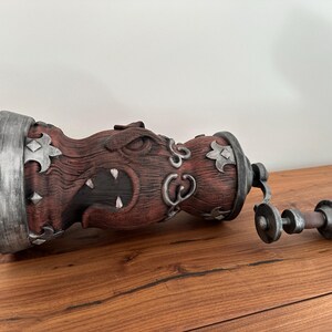 May include: A decorative, wooden-textured container with a menacing face carved into its side. The container is primarily reddish-brown with silver accents and a matching handle. The handle is connected to the container with a hinge.