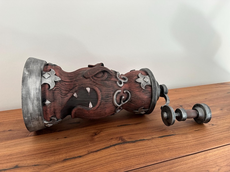 May include: A decorative, wooden-textured container with a menacing face carved into its side. The container is primarily reddish-brown with silver accents and a matching handle. The handle is connected to the container with a hinge.