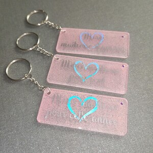 Peut inclure: Trois porte-cl&eacute;s rectangulaires roses avec des cha&icirc;nes et des anneaux argent&eacute;s. Chaque porte-cl&eacute;s pr&eacute;sente un motif de c&oelig;ur et du texte de couleur diff&eacute;rente. Le texte sur les porte-cl&eacute;s se lit "madre", "M" et "pour cette ann&eacute;e".