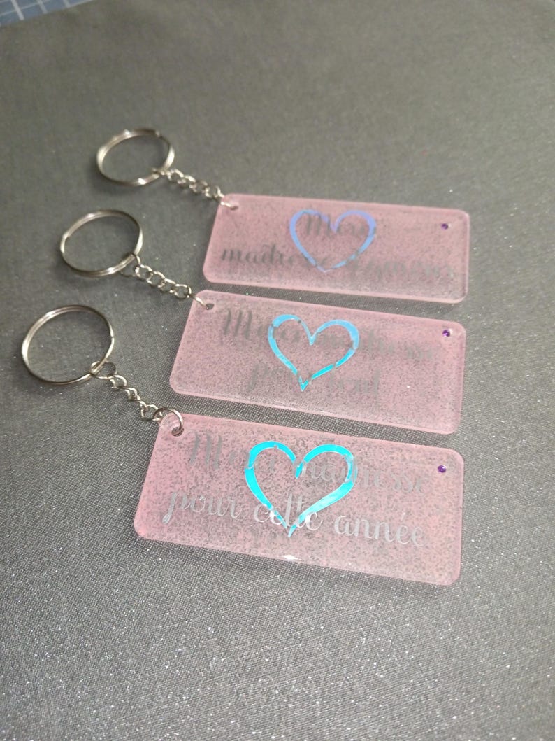 Peut inclure: Trois porte-cl&eacute;s rectangulaires roses avec des cha&icirc;nes et des anneaux argent&eacute;s. Chaque porte-cl&eacute;s pr&eacute;sente un motif de c&oelig;ur et du texte de couleur diff&eacute;rente. Le texte sur les porte-cl&eacute;s se lit "madre", "M" et "pour cette ann&eacute;e".